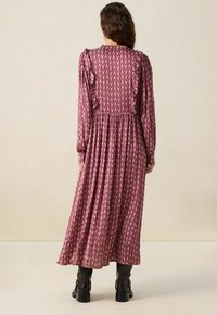 Une longue robe fluide avec un design de volants superposés, un imprimé géométrique violet et rose, des manches longues et une taille cintrée. Portée avec des bottes noires.