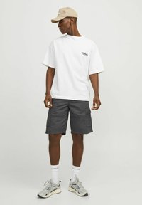 T-shirt oversize blanche avec logo sur la poitrine gauche, shorts cargo gris, casquette beige, baskets gris clair et chaussettes blanches avec texte noir.