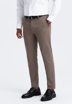 Uomo che indossa pantaloni beige a sigaretta, cintura nera, scarpe eleganti nere e camicia bianca a maniche lunghe con orologio al polso sinistro.