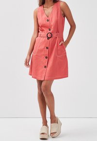 Robe sans manches corail avec un décolleté en V, des boutons à l'avant et des coutures apparentes. Elle est dotée d'une ceinture avec une boucle ronde et de poches latérales.