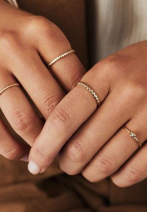 Goldene Ringe, die an Händen präsentiert werden; die Merkmale umfassen strukturierte, gedrehte und perlenbesetzte Designs, die in der Breite variieren und aufwendige Details aufweisen.