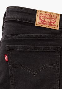 Black denim jeans featuring a tan leather logo patch with red and gold details. Stitching is in a lighter shade, with a red tab on the pocket.
