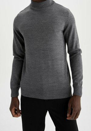Mann trägt einen engen grauen Rollkragenpullover mit gerippten Bündchen und Saum, kombiniert mit schwarzen Hosen vor einem schlichten Hintergrund.