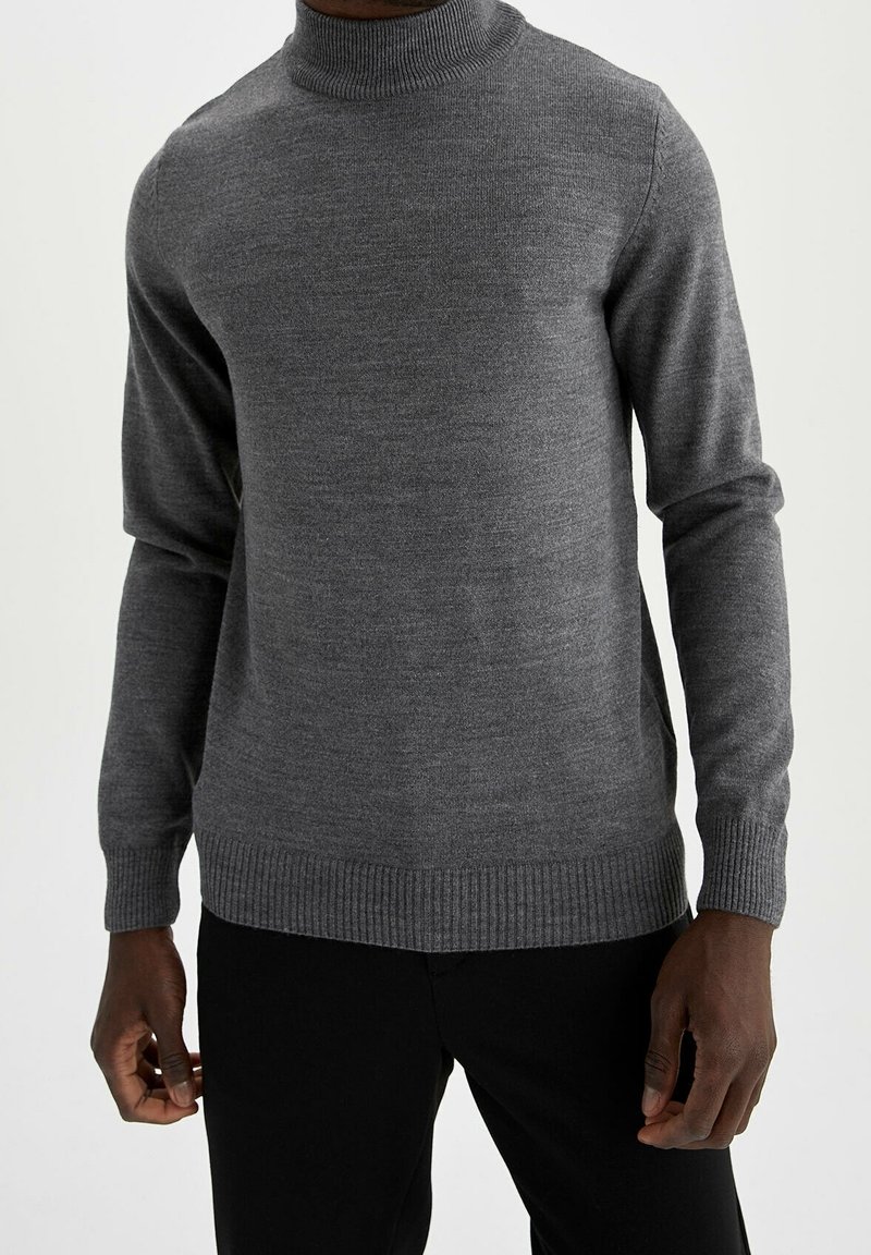 Homme portant un pull gris ajusté à col roulé avec poignets et ourlet côtelés, associé à un pantalon noir sur un fond uni.