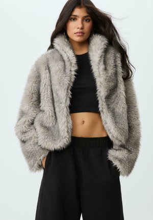 Junge Frau mit langen dunklen Haaren, die eine hellgraue Kunstfelljacke, ein schwarzes Crop-Top und schwarze Hosen trägt, steht mit den Händen in den Taschen.