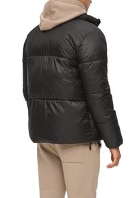 Schwarze Steppjacke mit Kapuze und gerippten Bündchen. Sie hat eine glatte, glänzende Außenseite und ein plüschiges Innenfutter und wird über beigen Hosen getragen.