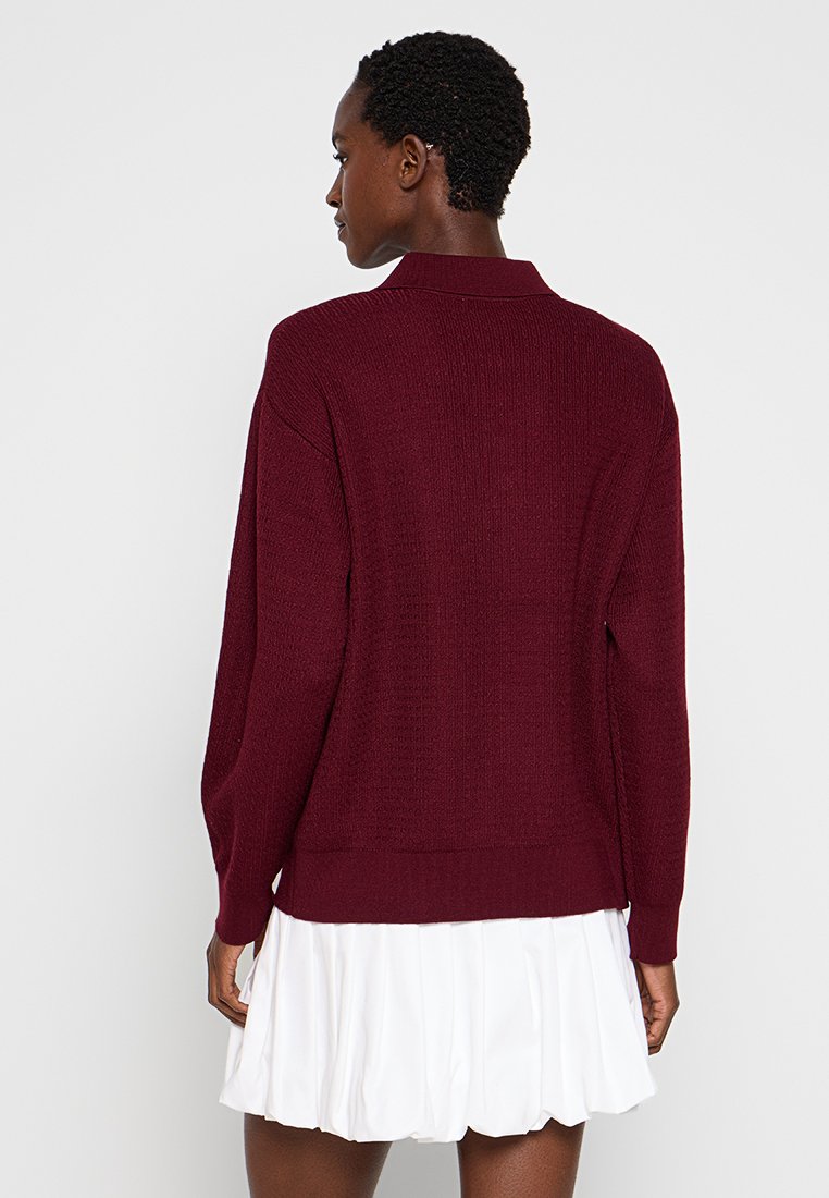 Maglione lavorato a maglia bordeaux con colletto, maniche lunghe e orlo a costine. Presenta un tessuto testurizzato e una vestibilità rilassata, abbinato a una gonna bianca plissettata.