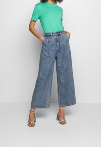 Vihreä lyhythihainen t-paita, korkeavyötäröiset leveälahkeiset siniset farkut ja musta-valkoiset raidalliset korkokengät nilkkaremmill ä.