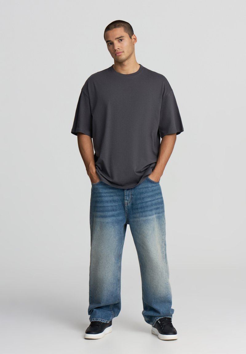 Czarnoszary, oversize t-shirt o luźnym kroju, zestawiony z jasnoniebieskimi spodniami o szerokich nogawkach z gradientowym wyblaknięciem. Czarne sneakersy dopełniają stylizację.