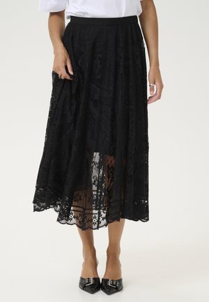Femme portant une jupe midi en dentelle noire avec des motifs floraux, associée à des chaussures noires à bout pointu et à un chemisier blanc.