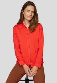 Red button-up shirt with a collar, long sleeves, and a smooth texture, paired with brown pants. Simple design with no visible patterns.