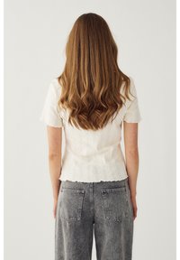 Frau mit langen, welligen braunen Haaren, die ein weißes, kurzärmeliges Strickoberteil und eine hoch taillierte graue Jeans trägt, von hinten vor einem einfarbigen Hintergrund.