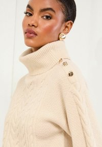 Béžový sveter s pletením a rebrovým rolákovým golierom, ozdobený kovovými gombíkmi na pleci. Mäkká textúra a uvoľnený strih.
