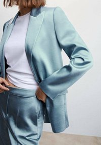Helle blaue satinierte Blazer mit breiten Revers und langen Ärmeln, kombiniert mit passenden satinierten Hosen. Das Model hält den Taillenbund und zeigt den Glanz des Stoffes.