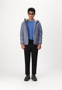 Giacca leggera grigia con fodera bianca, camicia blu, pantaloni neri e sneakers nere, in piedi su uno sfondo semplice.