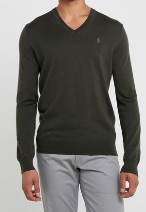 Homme portant un pull col V vert foncé avec un petit logo sur la poitrine et un pantalon gris clair, debout devant un fond uni.