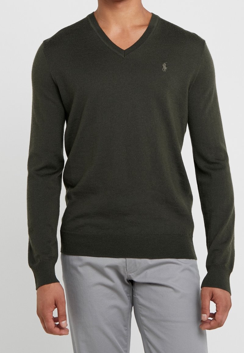Homme portant un pull col V vert foncé avec un petit logo sur la poitrine et un pantalon gris clair, debout devant un fond uni.