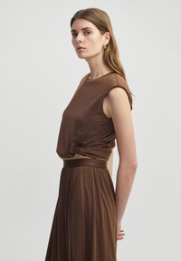 Bruin twee-delige outfit met een mouwloze top met geplooide details bij de taille en een lange, vloeiende rok. Zachte stofstructuur.