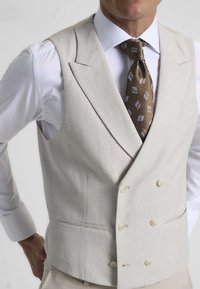 Chaleco de lino beige de doble botonadura con dos bolsillos frontales y botones claros. Lleva puesto sobre una camisa blanca de vestir, acompañado de una corbata marrón con patrón.