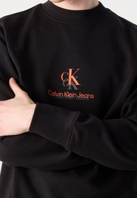 Černá mikina s pružným lemem na krku a manžetách. Má oranžové tištěné logo „CK“ a nápis „Calvin Klein Jeans“ v tučném písmu.