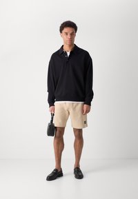 Svart tröja med krage, beiga shorts och svarta slip-on skor. Håller en liten svart väska. Har en avslappnad, lös design.