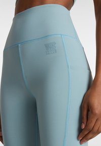 Leggings alta vita di colore azzurro chiaro con una texture liscia, caratterizzati da cuciture blu a contrasto e un logo Vans impresso nella cintura.