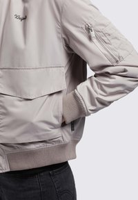 Svijetlosiva bomber jakna s prednjim džepom, džepom na ruci sa zatvaračem, rebrastim manžetama i glatkim materijalom s minimalnim detaljima šivanja.