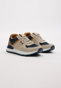 Zapatillas beige y marinas con materiales de gamuza y malla, con una media suela blanca y detalles en marrón. Cintas y un pequeño logo en el lateral.
