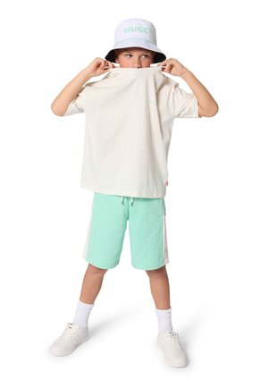 Kind draagt een pastelkleurige emmerhoed, witte oversized T-shirt opgetild naar de mond, mintgroene korte broek, witte sokken en witte sneakers, staat tegen een witte achtergrond.