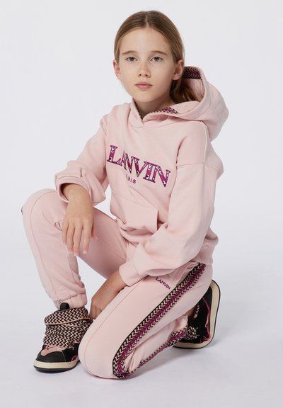 Ensemble en sweat à capuche et pantalon rose clair arborant le logo "LANVIN" en violet, avec des bandes latérales à motif tressé, complété par des chaussures noires et roses.