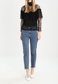 Kvinde iført en sort blonde-top med gennemsigtige ærmer, blå højtaljede jeans med frayed kanter og hvide spidse hæle, står foran en hvid baggrund.