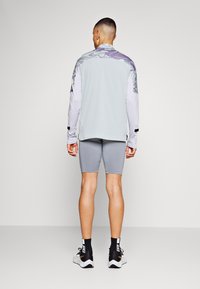 T-shirt de sport à manches longues gris clair avec une épaule à motif, associé à un short gris. Des baskets noir et blanc complètent la tenue.