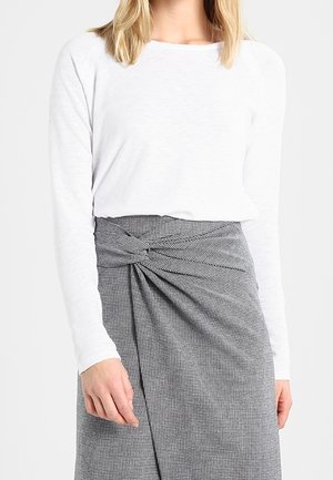 Femme portant une chemise blanche à manches longues et une jupe grise taille haute avec un détail de nœud torsadé à la taille.