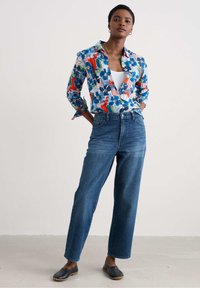 Chemise à bouton bleu à motifs floraux avec des accents rouges et verts, jeans taille haute bleu et chaussures noires à enfiler, présentés sur un fond neutre.