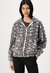 Veste en polaire à motifs noirs et blancs avec une fermeture éclair à l'avant, un col montant et des poignets élastiques. Présente un motif floral sur l'ensemble.