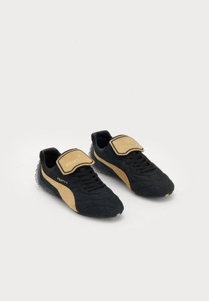 Par sorte og beige Puma Fenty sneakers med snører og store beige tungeflapper med Fenty-branding på en hvid baggrund.