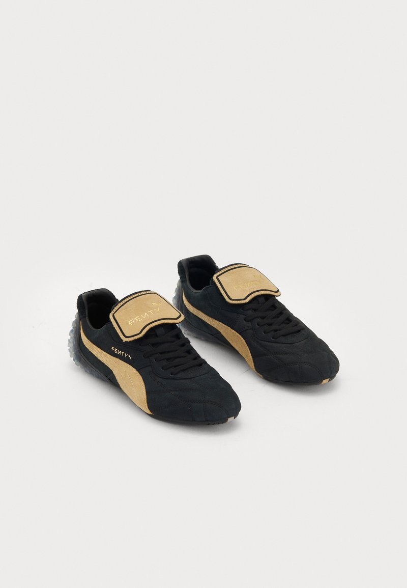 Paire de baskets Puma Fenty noires et beiges avec lacets et grandes languettes beiges arborant la marque Fenty sur fond blanc.