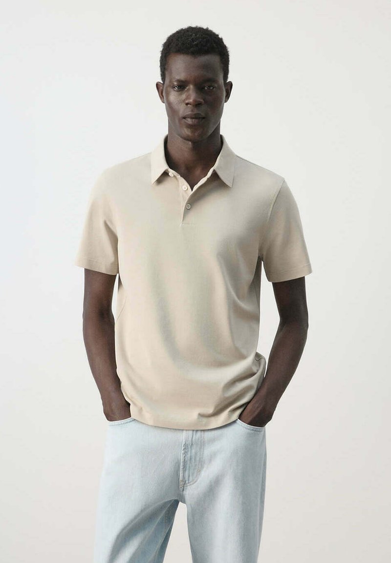 Hombre con camiseta tipo polo beige de manga corta y vaqueros azul claro, de pie con las manos en los bolsillos frente a un fondo claro y liso.