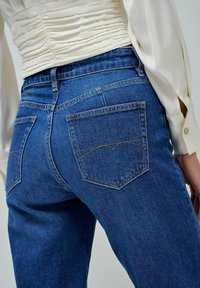 Los jeans de mezclilla azul tienen una cintura alta, dos bolsillos traseros con costura curva y una textura suave. Combinados con una blusa blanca con fruncidos.
