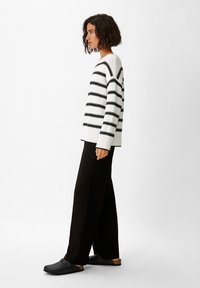 Gestreifter Pullover in Weiß und Schwarz, gestrickte Textur, entspannte Passform. Kombiniert mit weiten schwarzen Hosen und schwarzen Holzschuhen mit brauner Sohle.