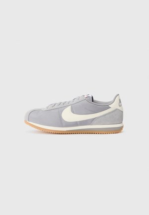 Las zapatillas de ante y nailon en gris presentan un prominente swoosh en crema de Nike, una suela de goma texturizada y un diseño con cordones. La suela de goma es de color marrón claro.