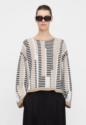 Donna che indossa un maglione oversize a righe beige, nere e bianche con maniche larghe e pantaloni neri, accessoriata con grandi occhiali da sole neri.