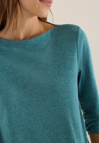 Tyrkysový dlouhý rukáv top s širokým výstřihem, vyrobený z měkké, texturované tkaniny. Má volný střih a decentní švy.
