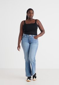 Top preto, jeans claros com corte flare e ténis pretos com solas brancas. A modelo está de pé num fundo simples, com as mãos nos bolsos.