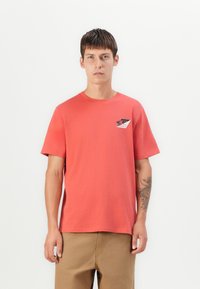 Young man with short brown hair wears coral red T-shirt with small black, white, and red graphic on chest, and light brown pants, against white wall.