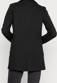 Cappotto nero in lana con trama a spina di pesce, caratterizzato da un colletto a bavero e tasche laterali. Taglio dritto con spacco posteriore.