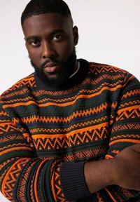Pull en tricot avec un motif multicolore comprenant des rayures orange, vert et marine. Les poignets côtelés et le col rond mettent en valeur le design.