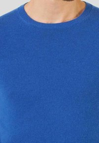 Närbild av en person som bär en enkel blå rundhalsad tröja, som visar halsen och övre delen av bröstet.