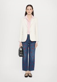 Blazer bianco sopra una camicia a righe rosa, abbinato a jeans a gamba larga blu con ricami floreali rossi e una borsa nera. Le scarpe nere completano il look.