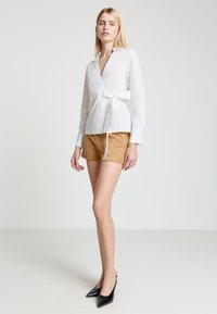 Weißes Hemd mit Knöpfen und Taillengürtel, kombiniert mit karamellfarbenen Shorts und schwarzen Slingback-Pumps mit spitzer Spitze. Schlichtes und maßgeschneidertes Design.